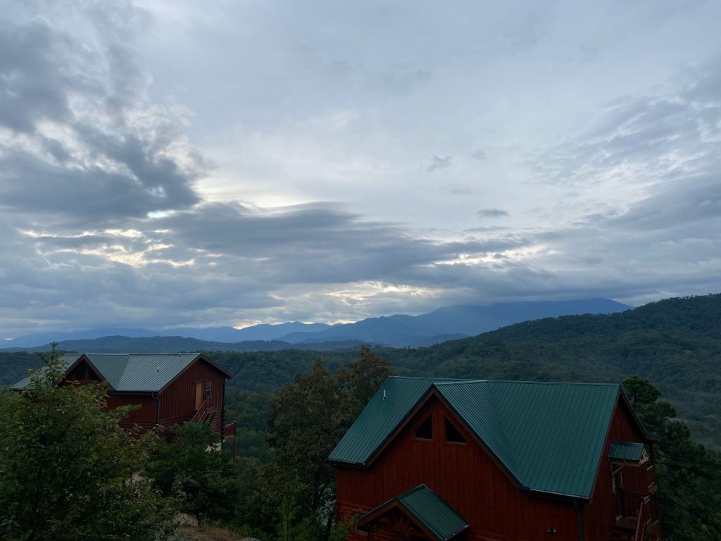 Two rustic wooden cabins with green metal roofs overlooking a lush, misty mountain range under a dramatic cloudy sky in smoky mountains