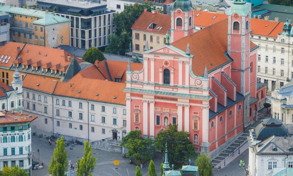 Elevated view of the pink Franciscan Church of the Annunciation with twin green domes in a historic Ljubljana city square.