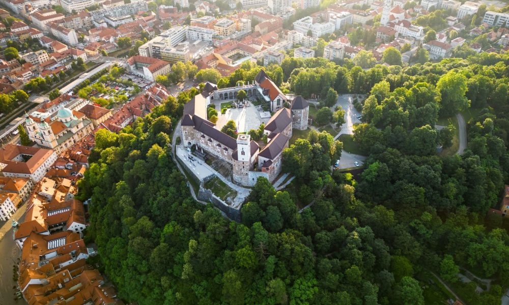 Aerial view of Ljubljana Castle atop a lush forested hill, surrounded by the historic terracotta-roofed city at golden hour.