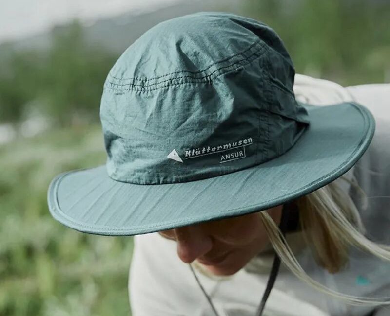 Teal Klättermusen Ansur wide-brimmed sun hat worn by a person looking down against a blurred green outdoor background.