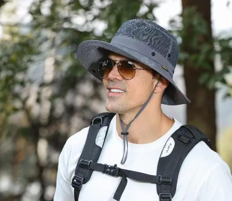 Smiling man wearing a grey mesh sun hat, aviator sunglasses, and a black backpack outdoors against a blurred forest background.
