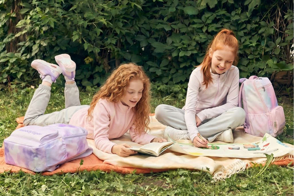 Two young girls reading and drawing on a grassy outdoor picnic blanket, accompanied by pastel tie-dye moodytiger backpacks.