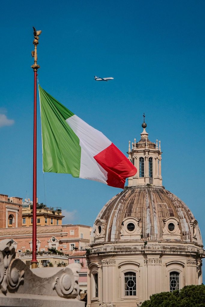 Italian flag waving in the wind beside a historic domed church in Rome, with an airplane flying in the clear blue sky.