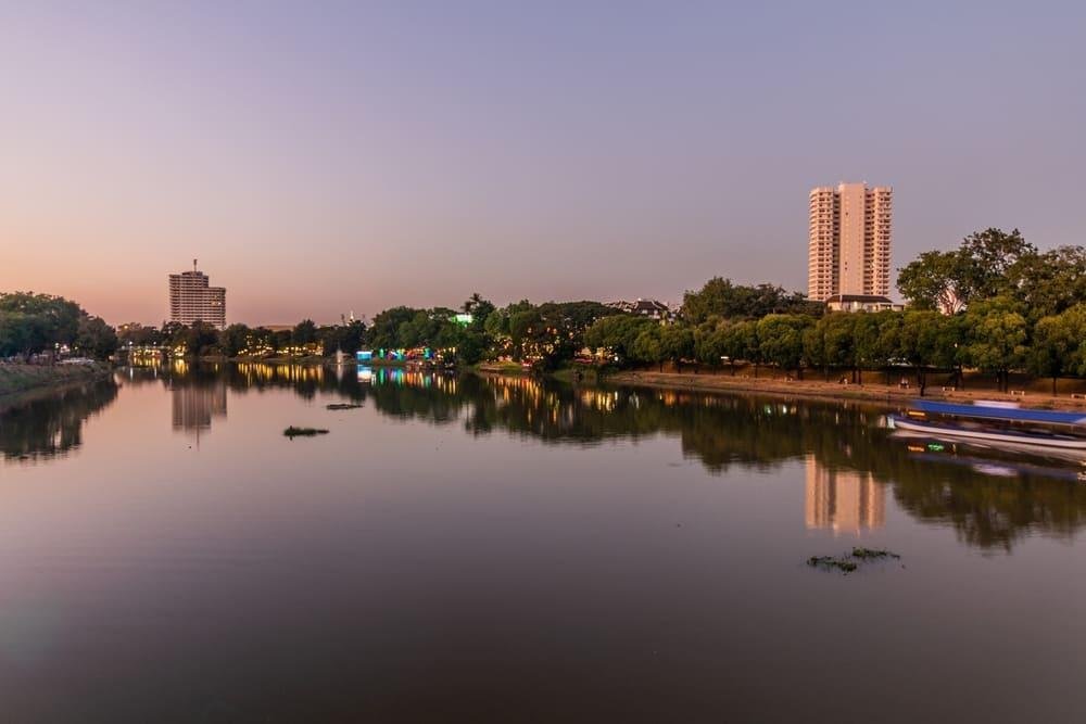 Calm urban river at twilight reflecting a pastel sky, tree-lined banks, and city buildings, with a blurred boat moving past in Ping River in Chiang Mai