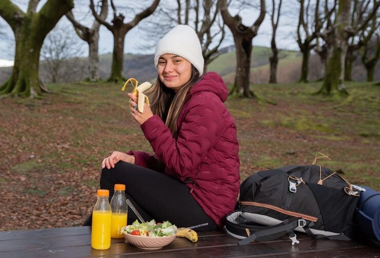 Smiling woman in a maroon jacket and beanie eating a banana at a picnic table with juice and salad during a hike.