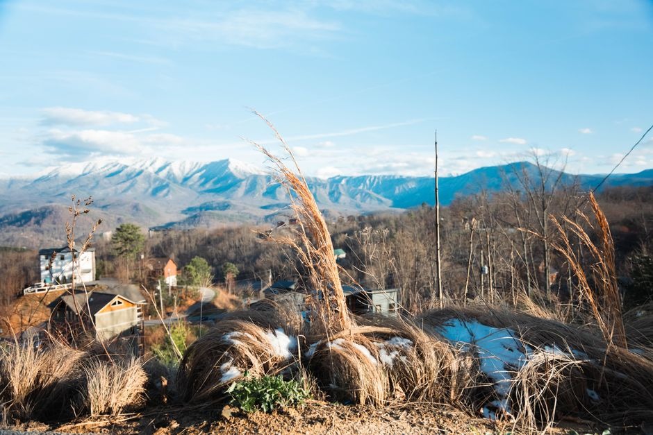 Winter mountain landscape with dry grass and snow patches overlooking valley cabins and snow-capped peaks under a blue sky in Gatlinburg