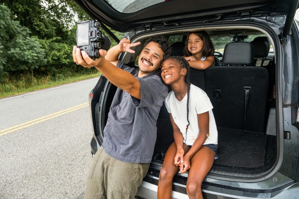 A smiling man and two young girls taking a happy road trip selfie with a digital camera at the open trunk of an SUV.