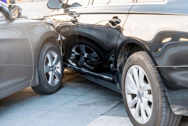 Close-up of a silver car's front bumper crashed into the dented side door of a black car on a paved street in daylight.