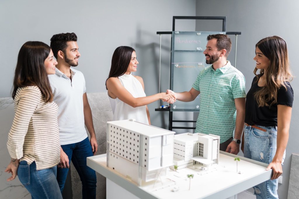 Smiling professionals shake hands over a white architectural building model during a meeting in a modern design office.