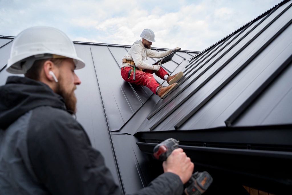 Two construction workers in white hard hats installing dark grey standing seam metal roofing panels on an overcast day.