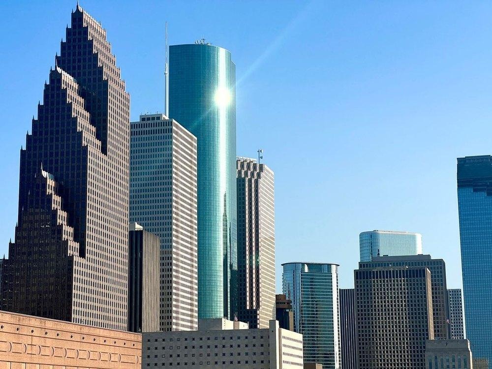 Downtown Houston skyline on a clear day, featuring a bright sun glare reflecting off a tall, cylindrical blue glass skyscraper.