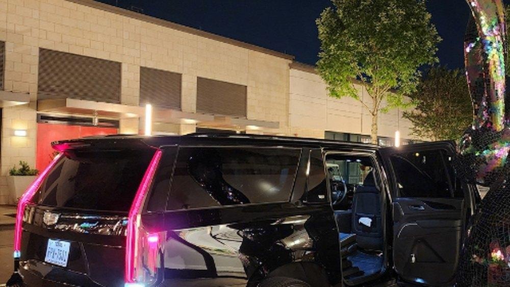 Black luxury SUV parked at night with its rear passenger door open and glowing red taillights outside a lit building.