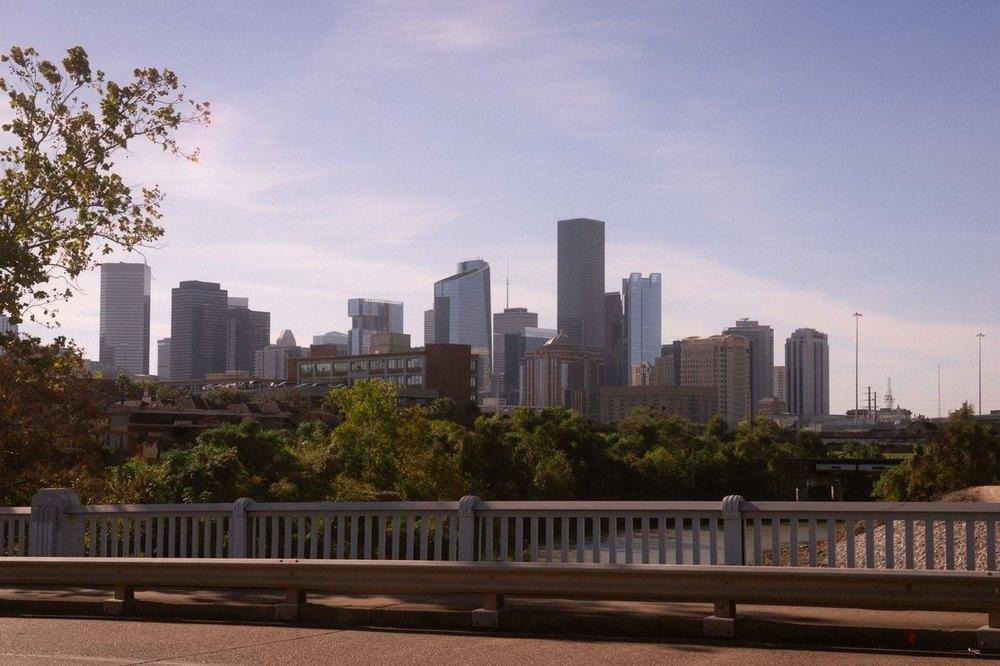 Houston downtown skyline rising above lush green trees, viewed from behind a concrete bridge railing under a hazy blue sky.