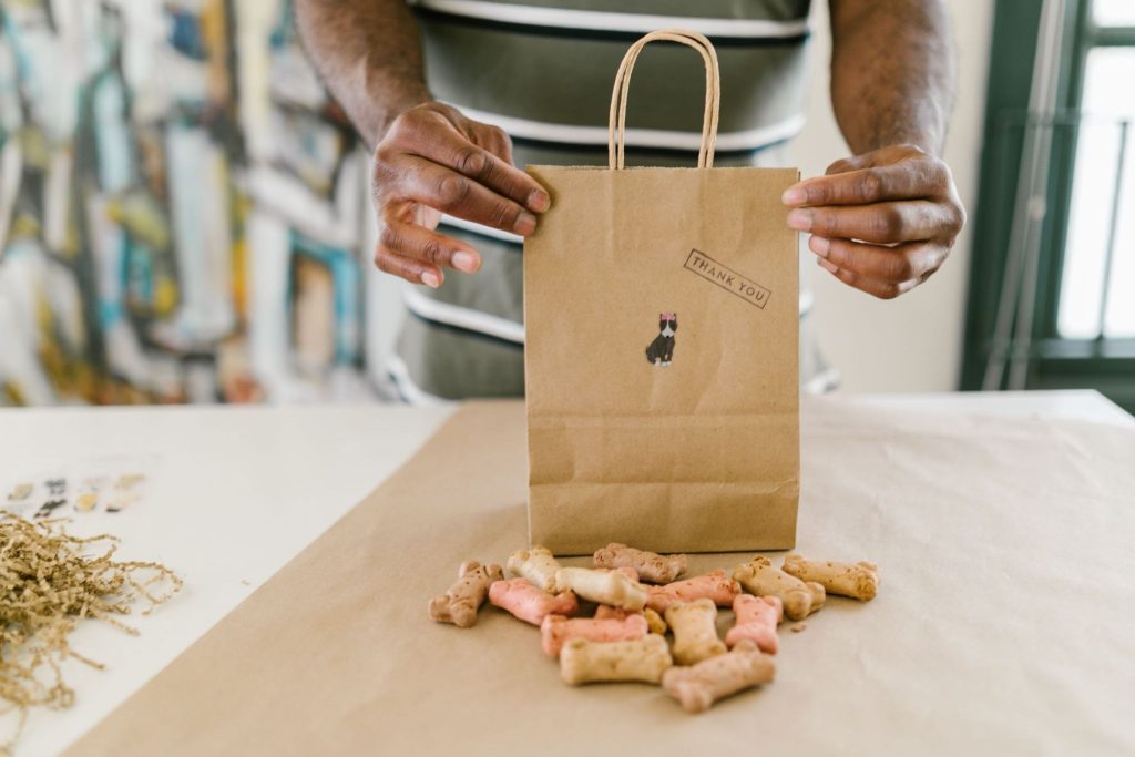 Person holding a brown paper gift bag stamped with "Thank You" behind a pile of bone-shaped dog treats on a craft table.