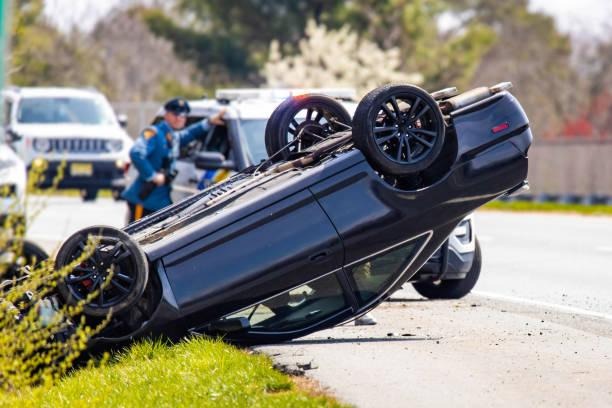 Upside-down black car on a roadside after a crash, with a police officer and police vehicle in the blurred background.