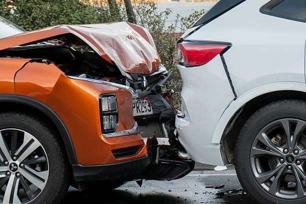 Close-up of a rear-end collision between an orange SUV with a heavily crumpled hood and a white SUV on an asphalt road.