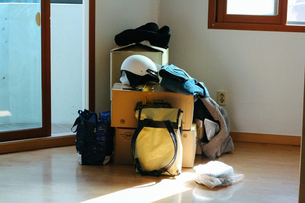 Cardboard moving boxes piled with a white helmet, yellow backpack, and clothes in the sunlit corner of an empty room.