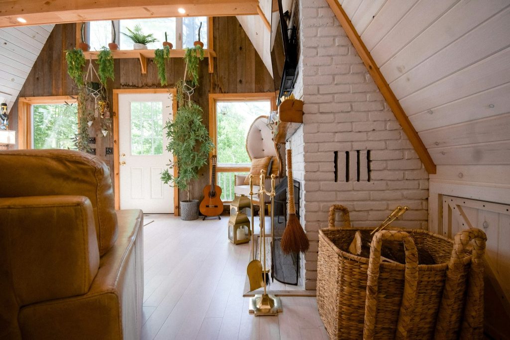 Cozy A-frame cabin interior featuring a white brick fireplace, rustic wood paneling, an acoustic guitar, and lush potted plants.