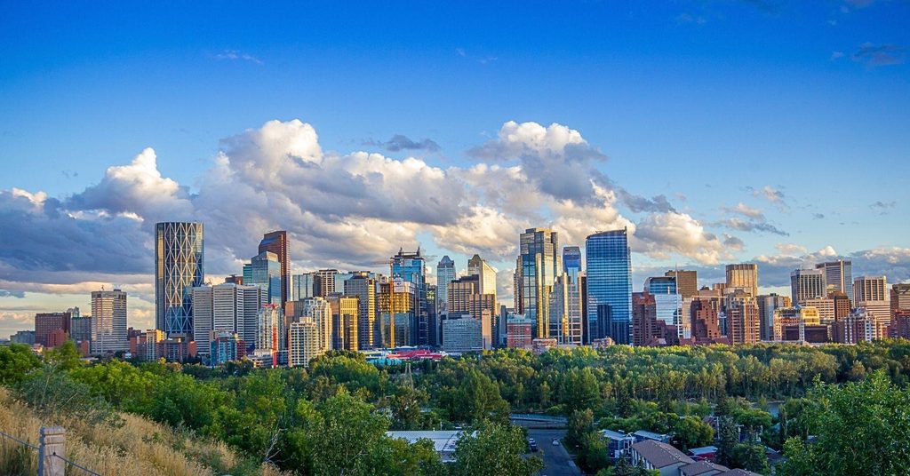 Modern city skyline lit by golden hour sunlight, towering over a lush green forest canopy beneath a bright, cloudy blue sky in Calgary