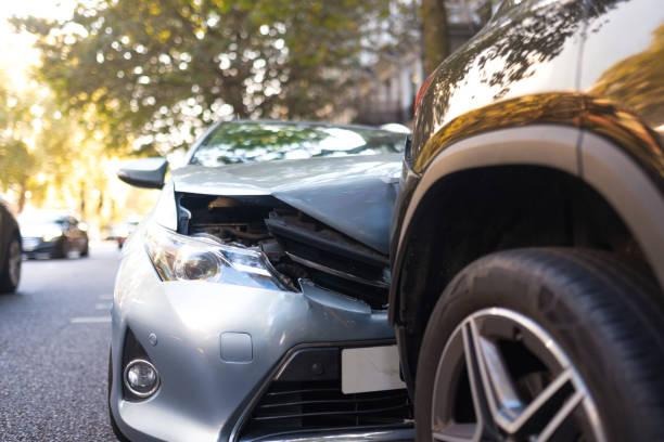 Close-up of a silver car with a heavily crumpled hood after rear-ending a dark vehicle on a sunny, tree-lined city street.