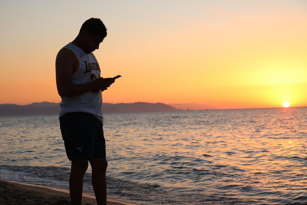 Man in a basketball jersey standing on a beach at sunset, looking down at his smartphone beside the ocean.   