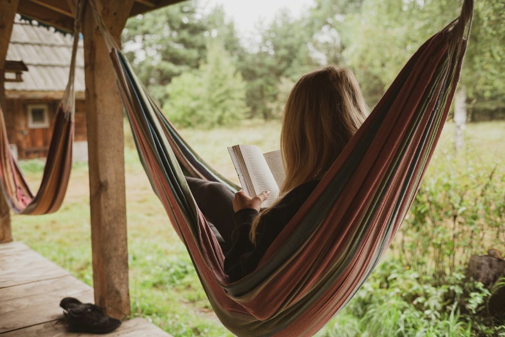 Woman with blonde hair relaxing in a striped hammock on a rustic wooden porch, reading a book against a green forest background.