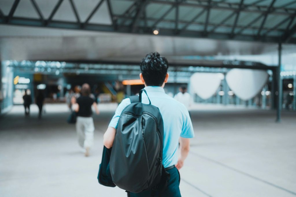 Back view of a man carrying a black backpack while walking through a modern, blurred urban transit concourse.