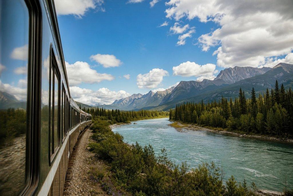Passenger train traveling beside a turquoise river and dense evergreen forest towards rugged mountain peaks under a blue sky.