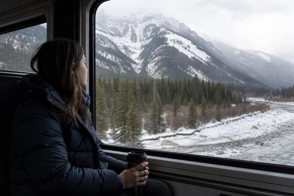 Woman in a puffer jacket looking out a train window at a snow-covered mountain, evergreen forest, and frozen river.