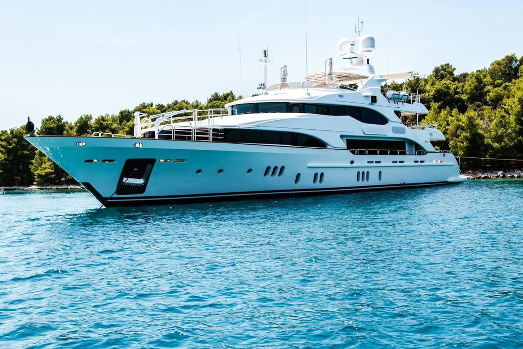 Large white luxury superyacht anchored in bright turquoise water near a lush green, tree-lined coast on a sunny day.