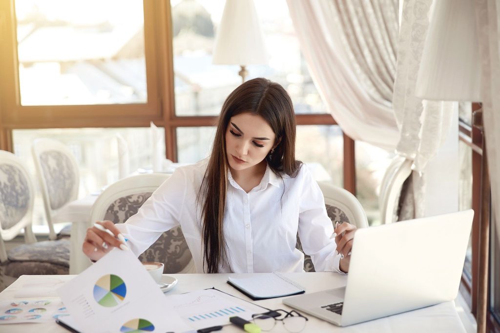 Young professional woman in a white shirt analyzing printed business charts and working on a laptop in a bright, sunny cafe.