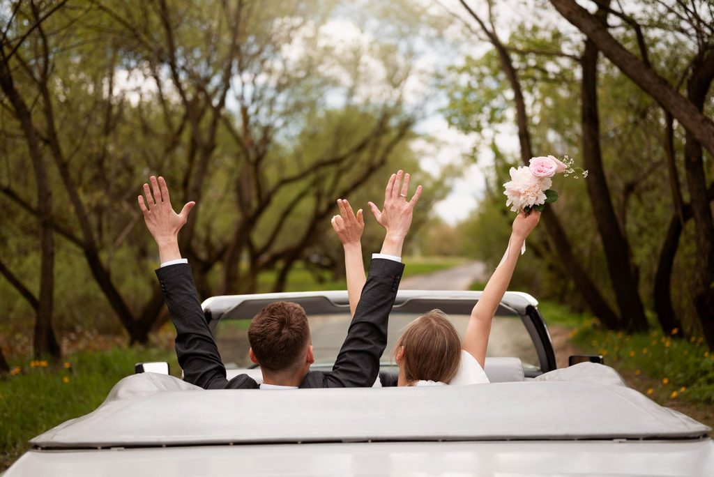 Newlywed couple in a white convertible driving down a tree-lined country road, raising their arms in joyful celebration.