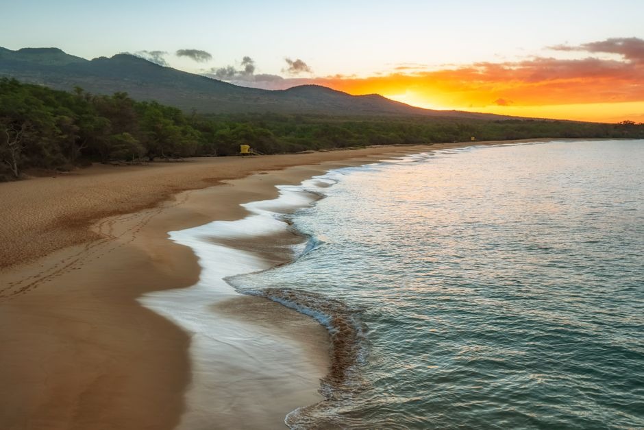 Serene tropical beach at sunset with gentle ocean waves, golden sand, lush green trees, and distant rolling mountains in Maui