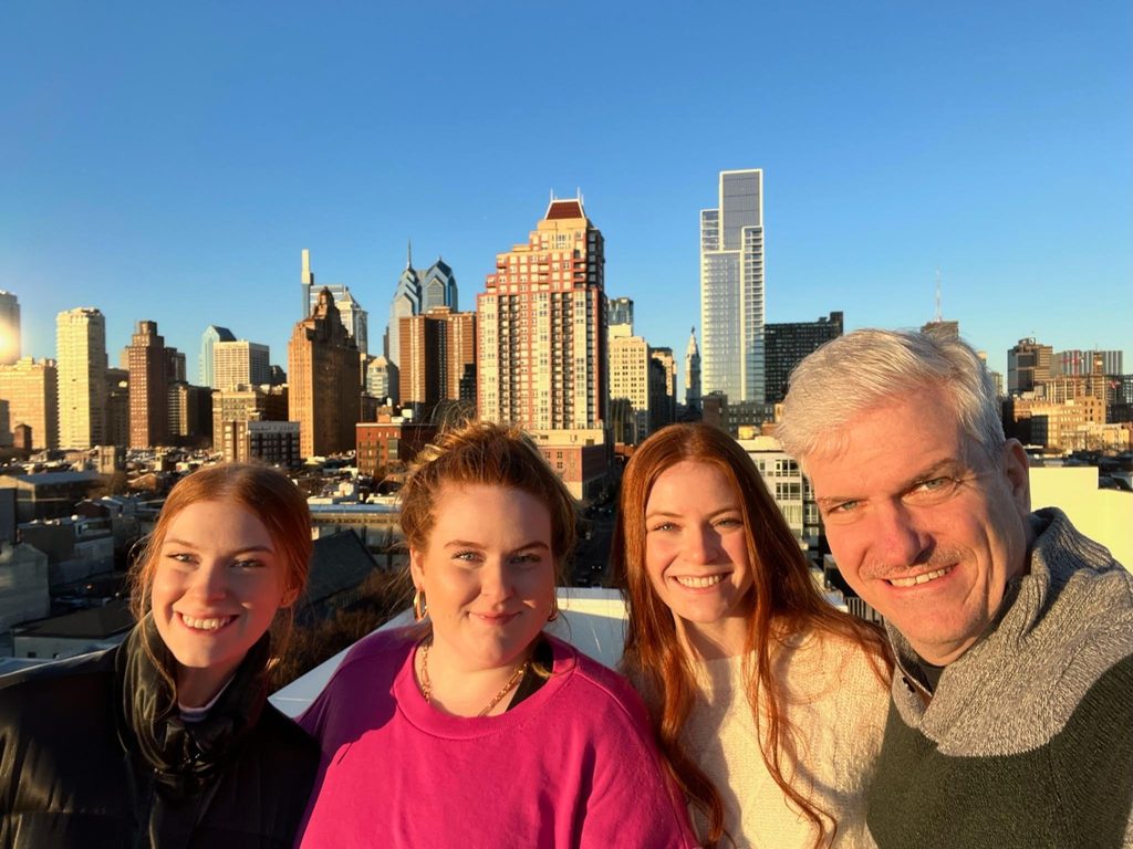 Group selfie of four smiling people on a rooftop during golden hour, with the Philadelphia skyline in the background.   
