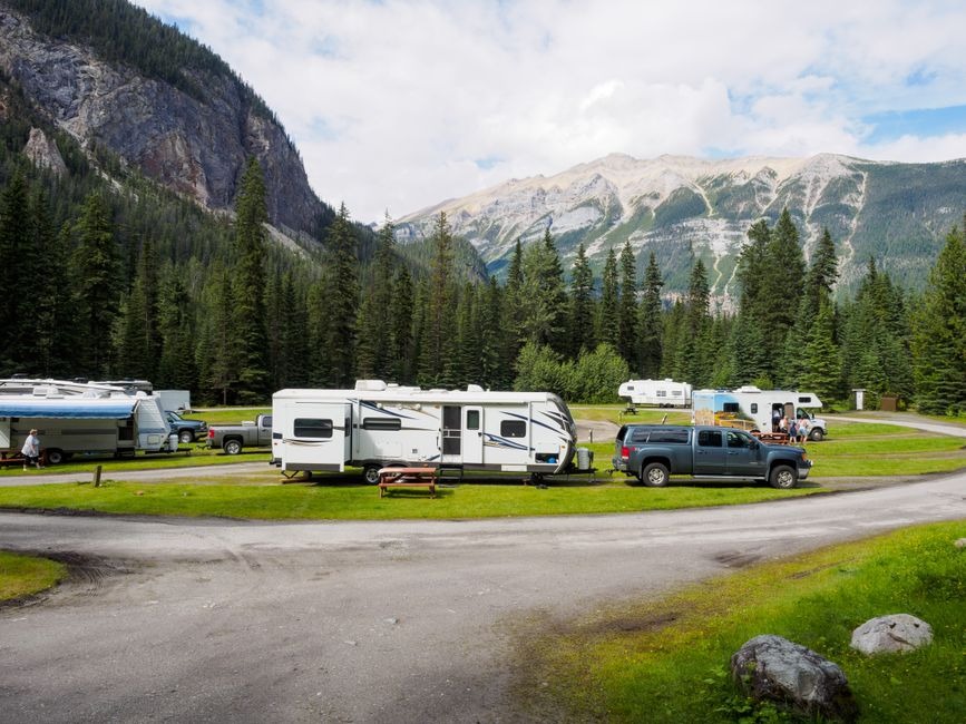 White RVs and trucks parked in a grassy campground surrounded by dense pine forests and towering mountain peaks in Kicking Horse Campground in Yoho National Park, British Columbia, Canada