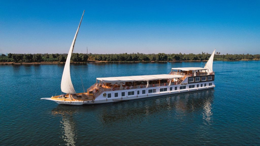 Traditional white dahabiya riverboat with a large lateen sail cruising on a calm blue river alongside a lush green shoreline.