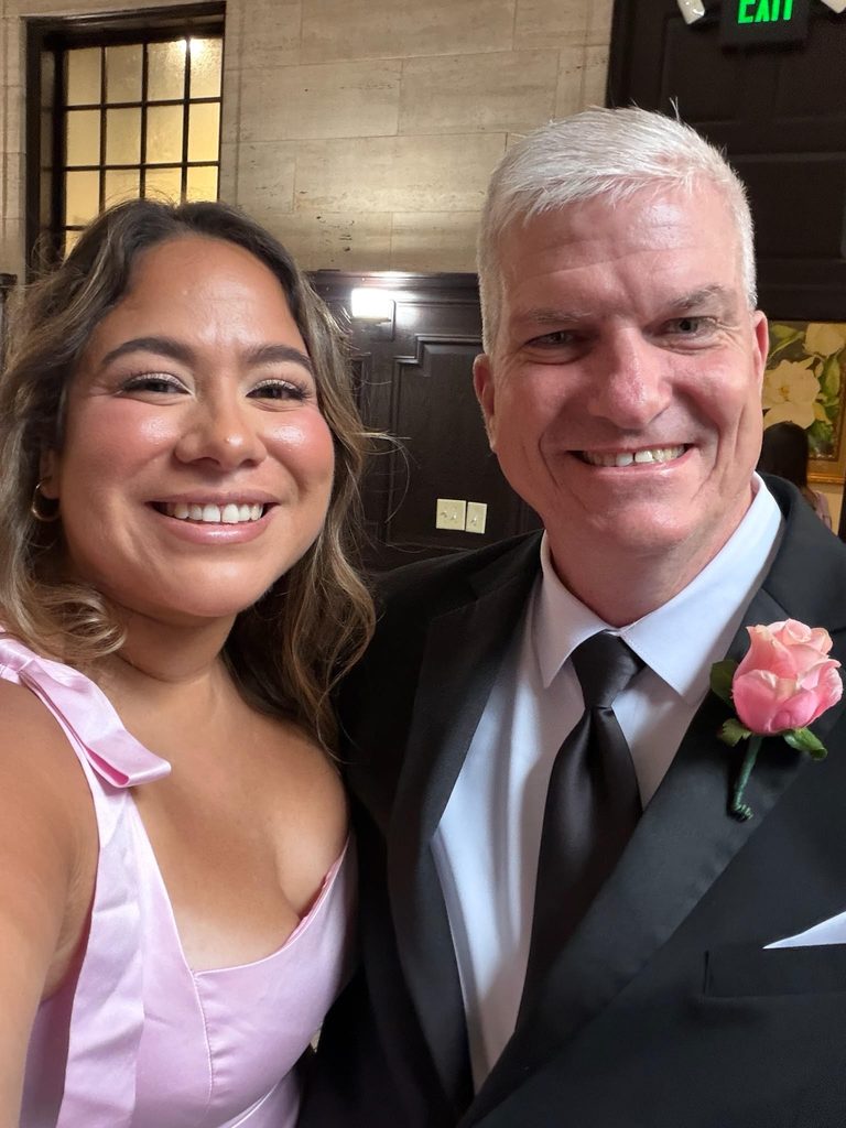 Selfie of a smiling woman in a pink dress and a man in a black suit with a pink rose boutonniere at a formal event.   