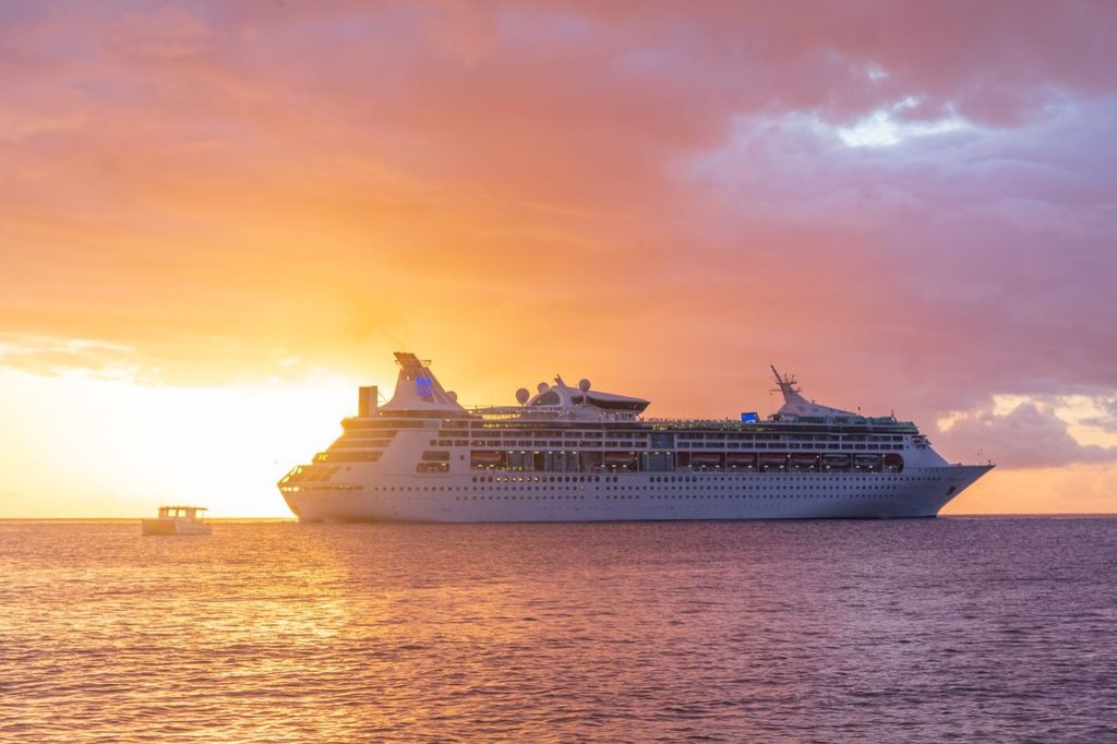 Large white cruise ship and a smaller boat sailing on the ocean under a vibrant orange, pink, and purple sunset.   