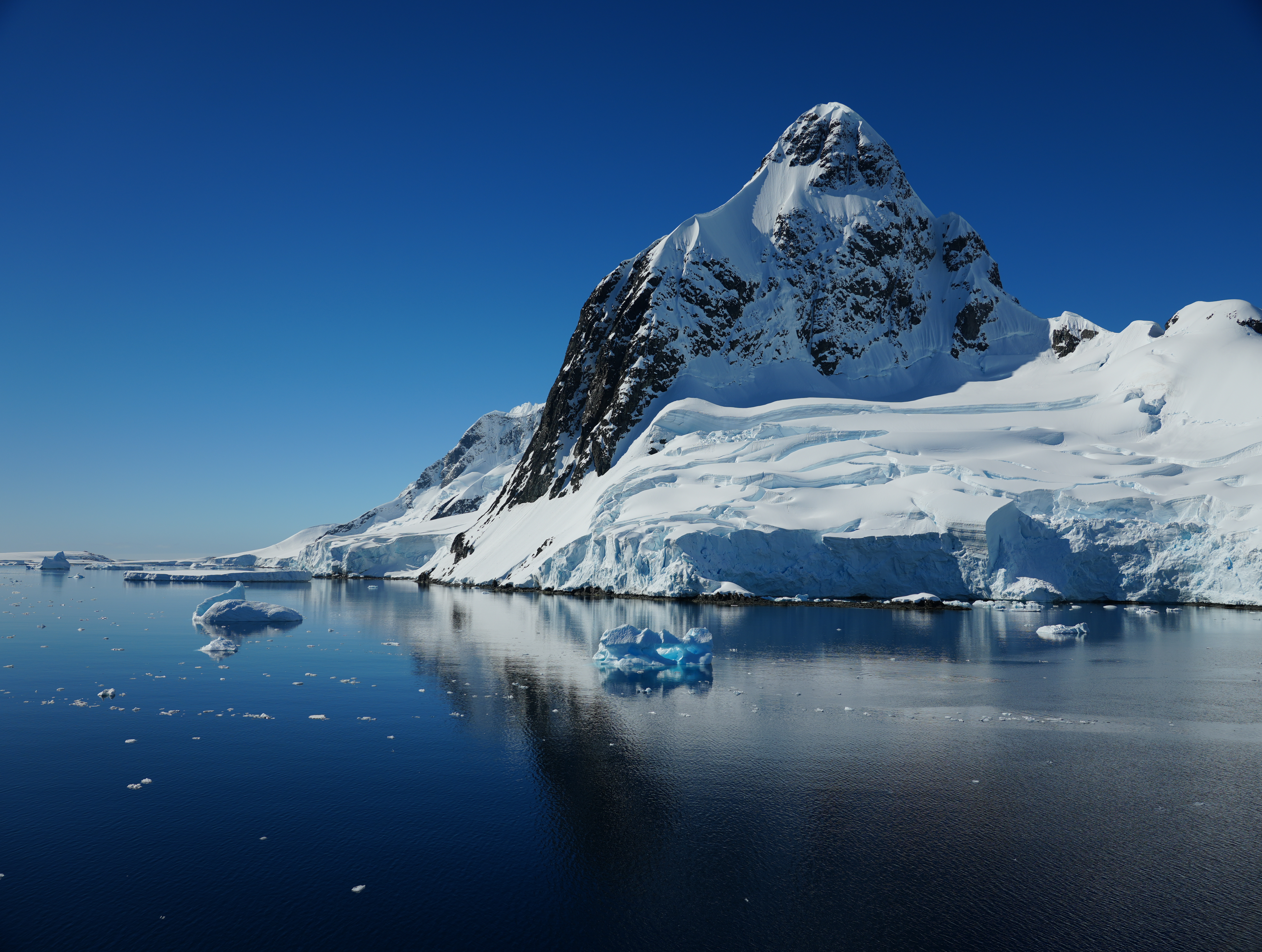 A majestic snow-capped mountain rises against a clear blue sky, surrounded by calm waters with floating icebergs in the foreground.