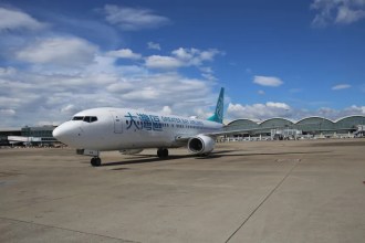 A Greater Bay Airlines aircraft is parked on the tarmac at an airport with a clear blue sky and scattered clouds in the background. The terminal building with its distinctive curved roof design is visible behind the plane.