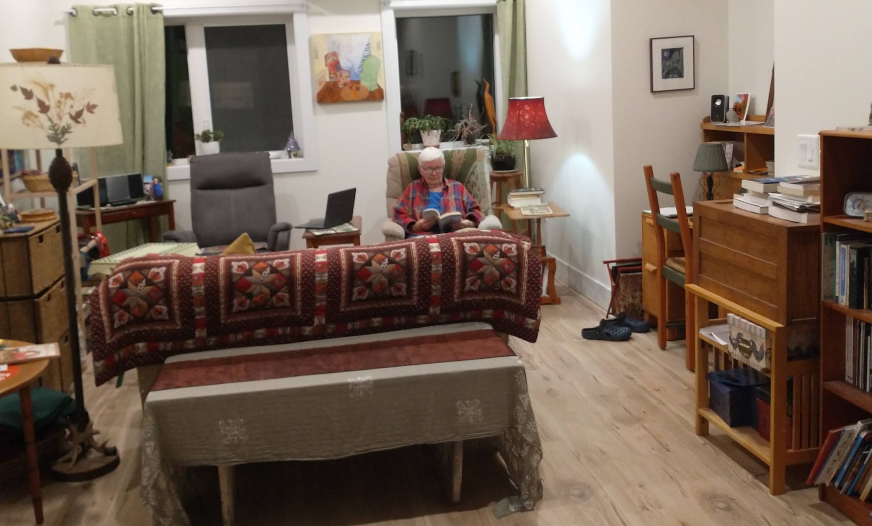 An older person wearing glasses and a red plaid shirt reads in an armchair in a warmly furnished living room. A patterned quilt is draped over the sofa in the foreground, with books and lamps around the room.
