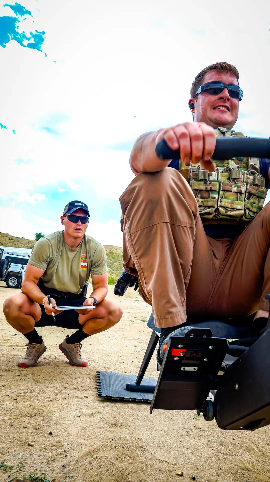 Man wearing tactical gear uses a rowing machine outdoors while a coach observes and takes notes.