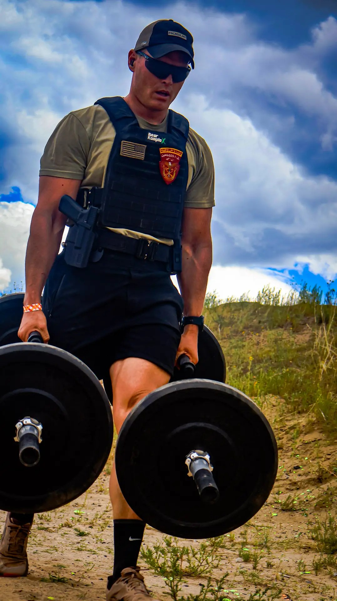 Man performing a farmer’s carry with barbells outdoors while wearing a tactical vest and gear.