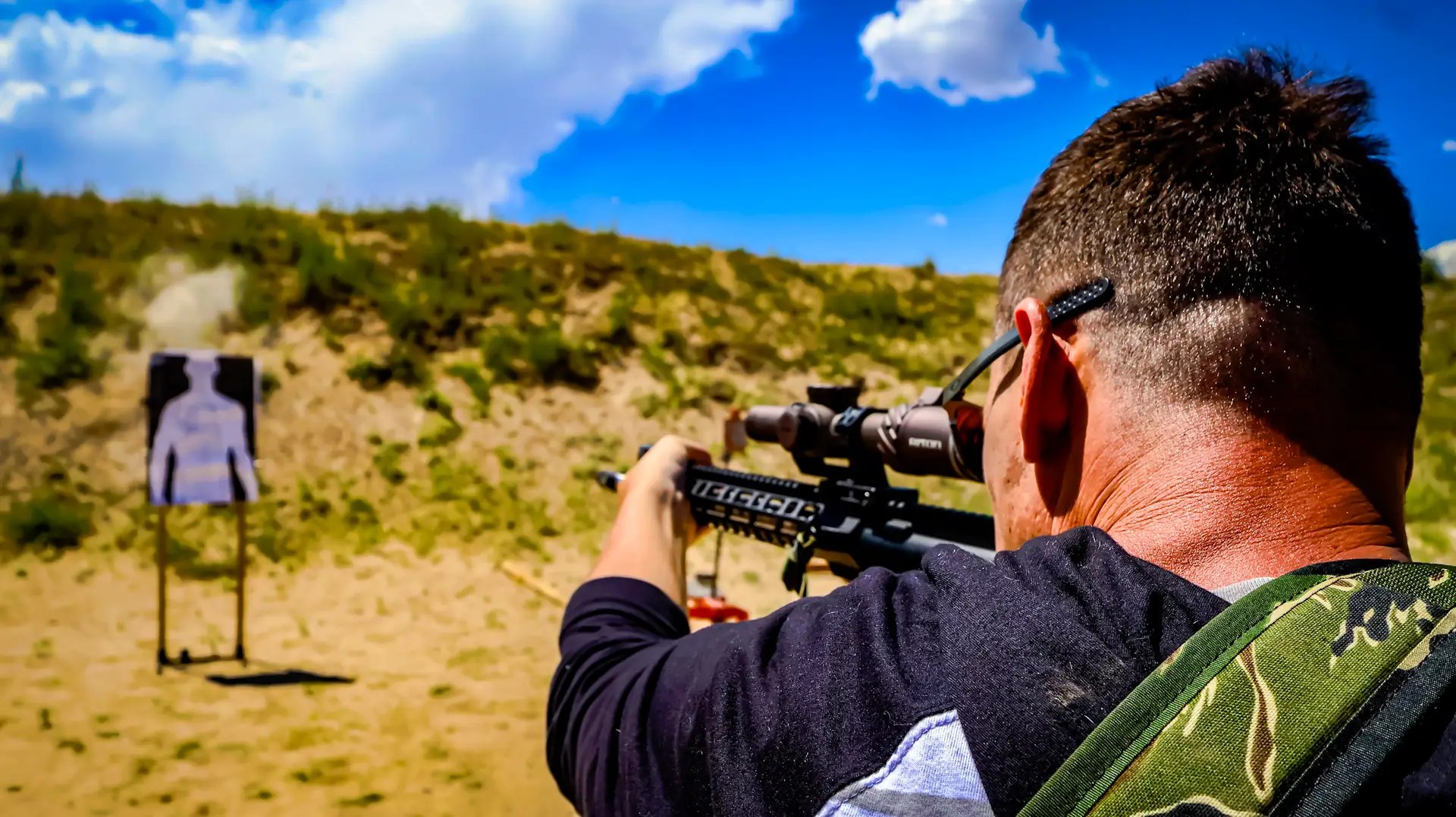 Shooter aiming a scoped rifle at a silhouette target during live fire at an outdoor range under a bright blue sky.