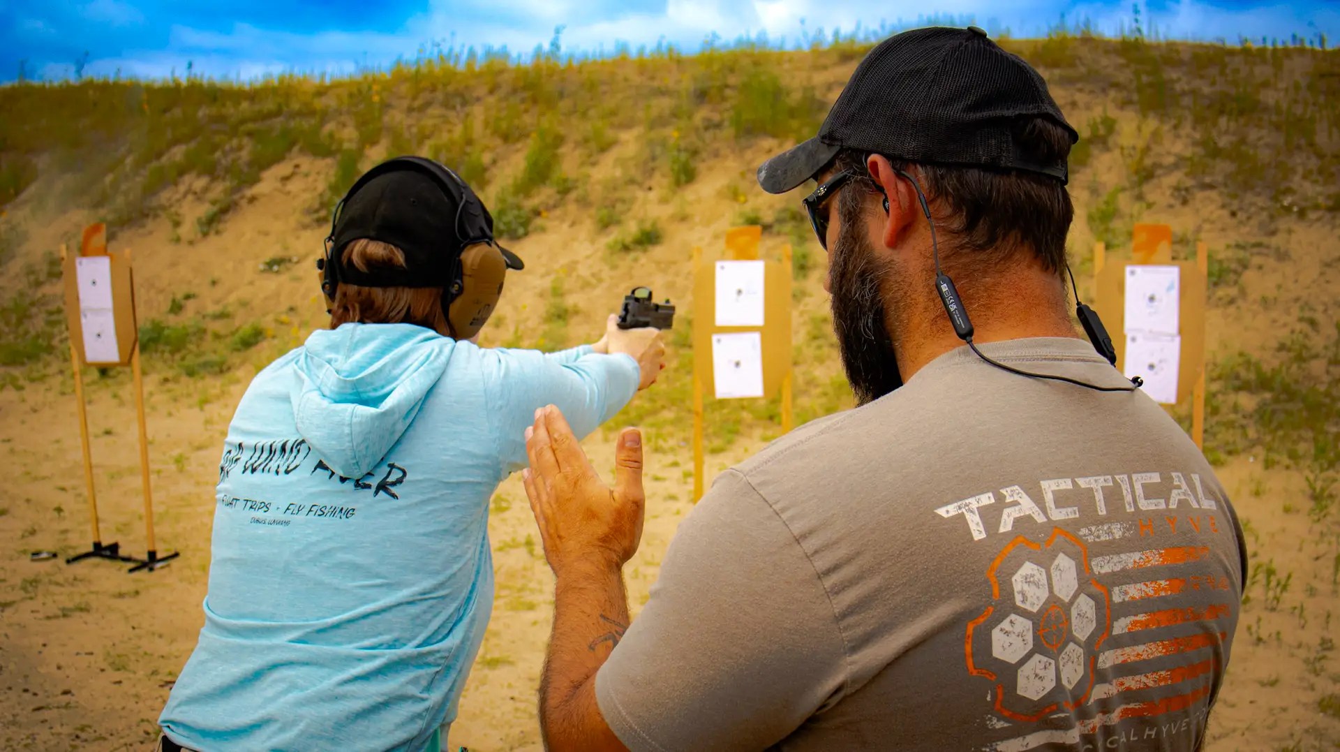 Firearms instructor coaching a student on pistol shooting at an outdoor range with paper targets in the background.