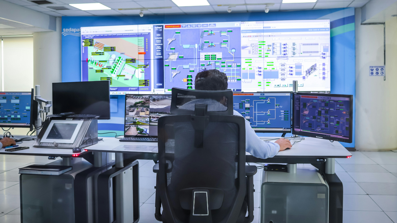 Operator seated with back to camera at a control desk, monitoring multiple computer screens and large wall displays with system diagrams.