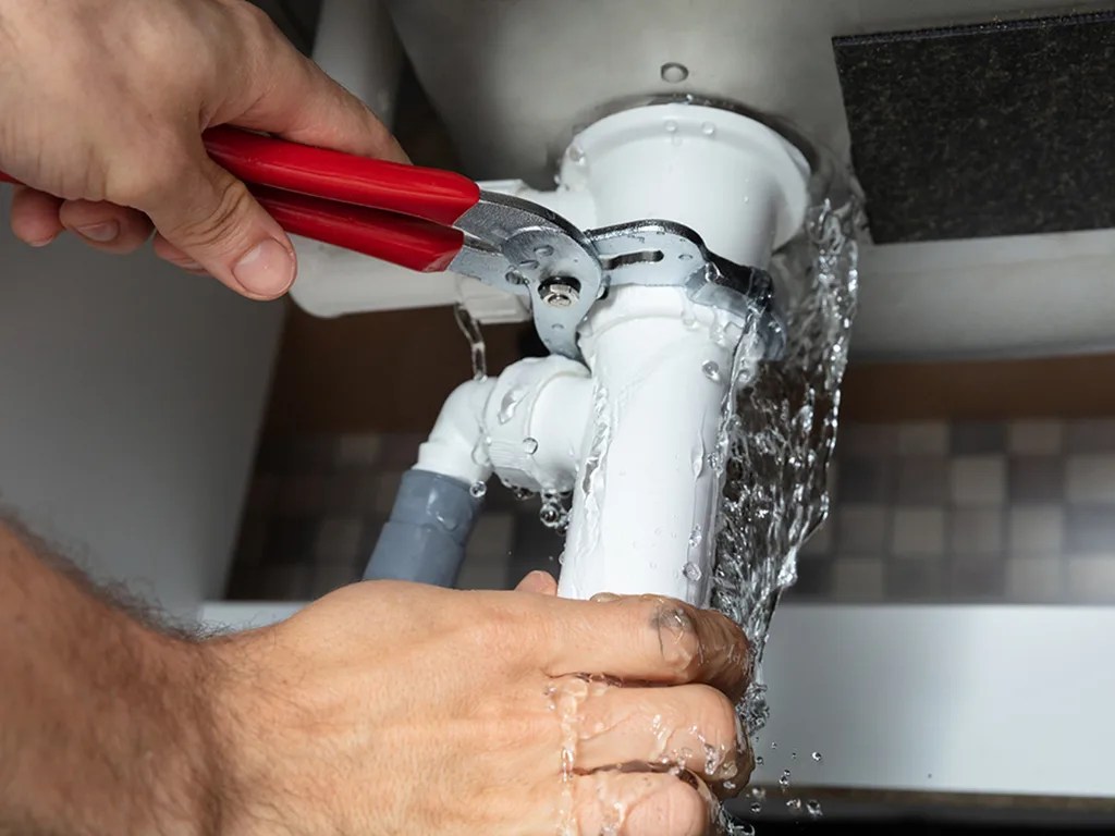 Emergency plumbing services stopping an active leak under a sink with channel‑lock pliers