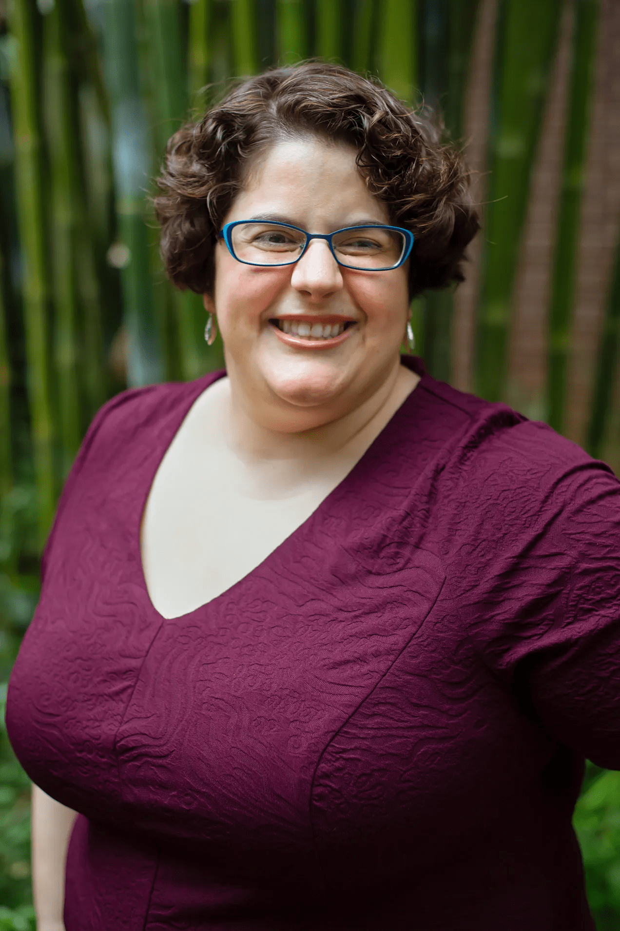 Emily K. Michael, smiling, short dark curly hair and blue-framed glasses, wearing a burgundy top, standing outdoors in front of bamboo.