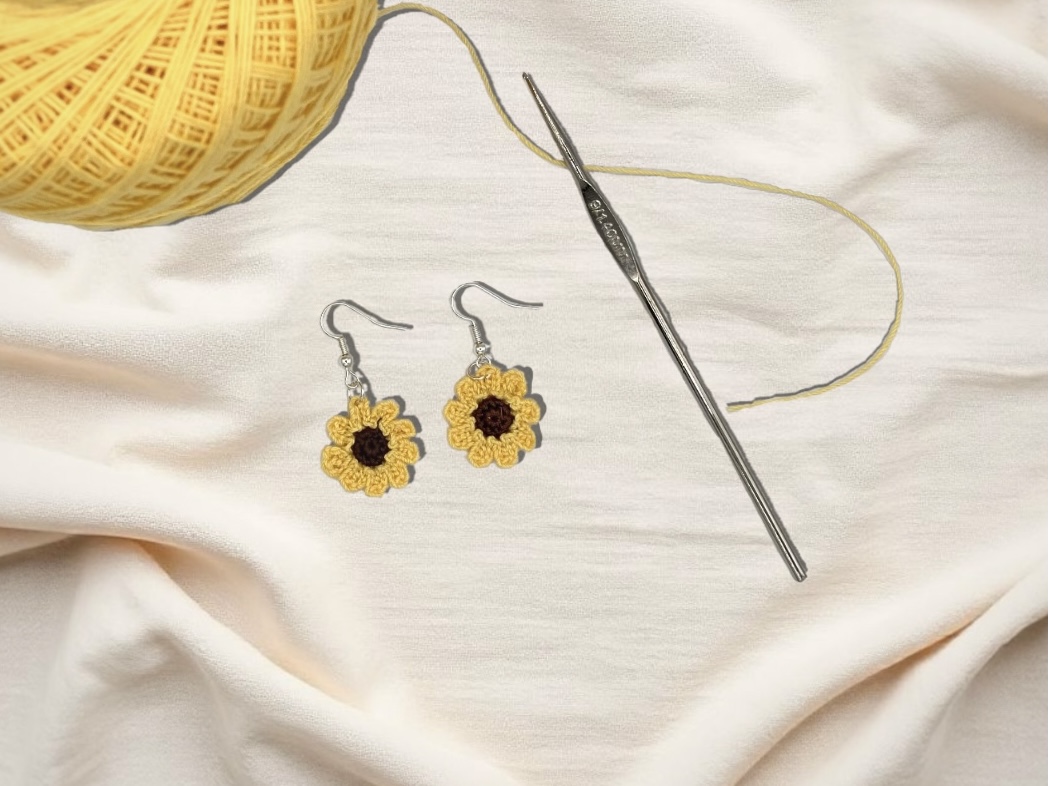 Crochet sunflower earrings, laying on a cloth, with a yellow ball of crochet thread, and metal crochet hooks