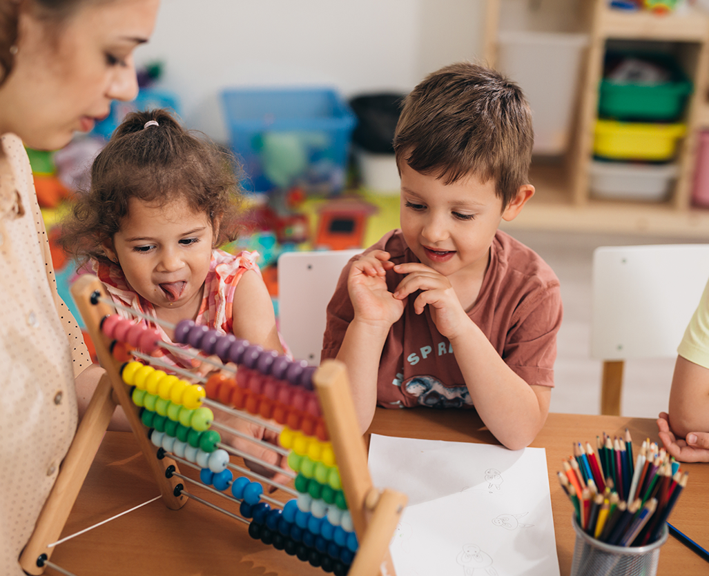 Una maestra de preescolar y dos niños pequeños están sentados en una mesa jugando con un ábaco colorido, con hojas para dibujar y lápices de colores cerca.